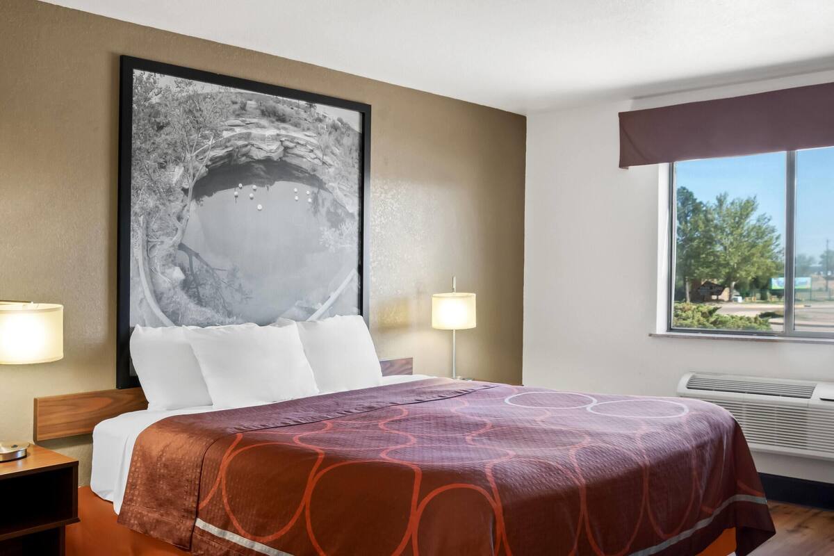 Ein Hotelzimmer mit einem großen Bett, zwei Nachttischlampen, einem gerahmten Bild an der Wand und einem Fenster mit Blick auf Bäume.