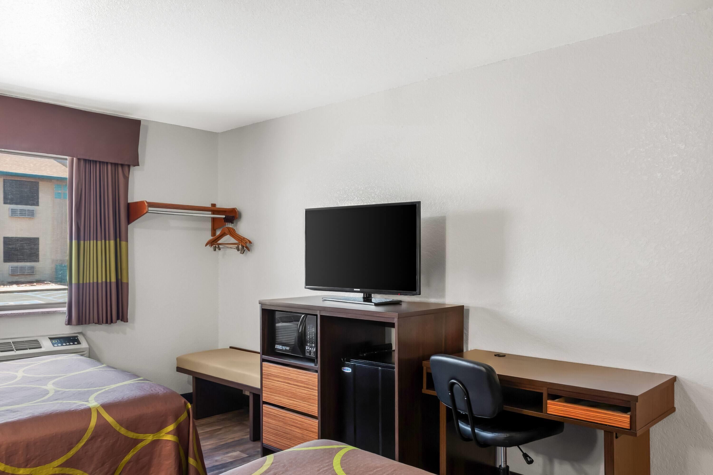 A hotel room with a bed, a desk, a chair, a television, a mini-fridge, and a window with curtains.