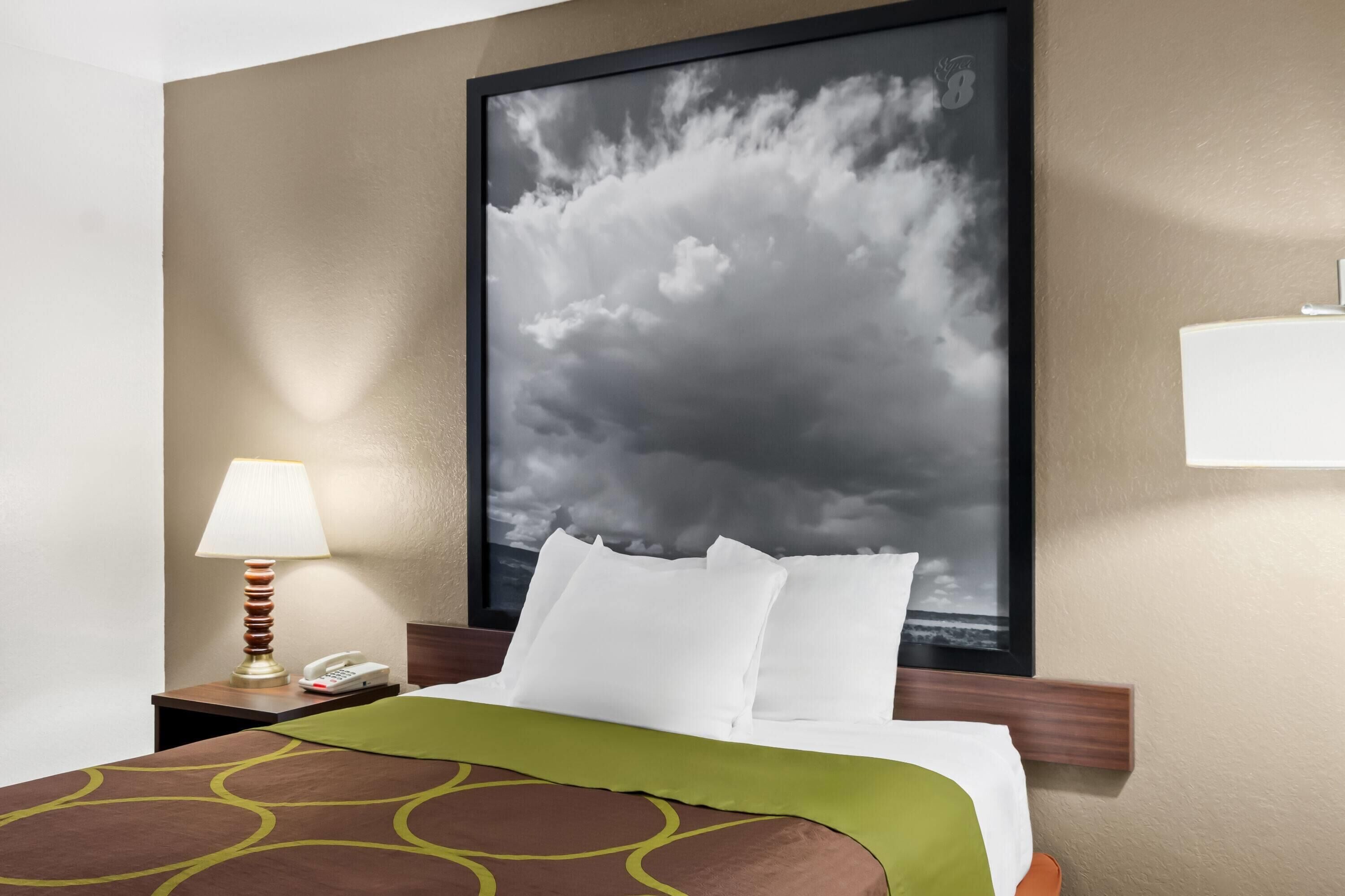 A hotel room with a bed, a nightstand with a lamp, a phone, and a framed picture of clouds on the wall.