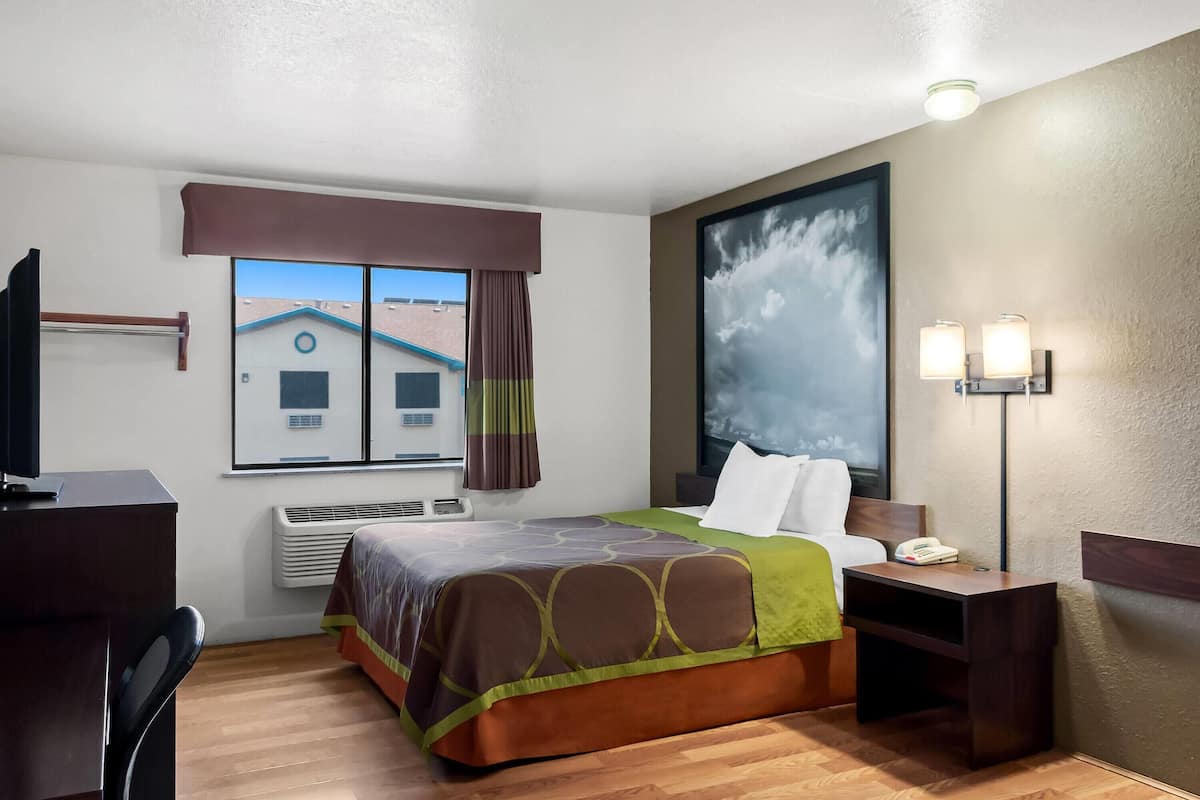 Ein Hotelzimmer mit Bett, Schreibtisch, Stuhl, Nachttisch, Lampe, einem Fenster mit Vorhängen, einer Klimaanlage und einem Bild an der Wand.