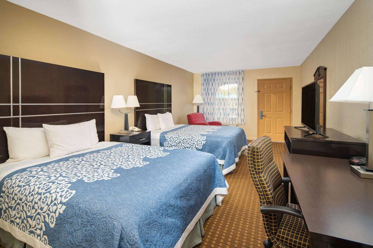 Ein Hotelzimmer mit zwei Betten, einem Schreibtisch, einem Fernseher und einem Fenster mit gemusterten Vorhängen.