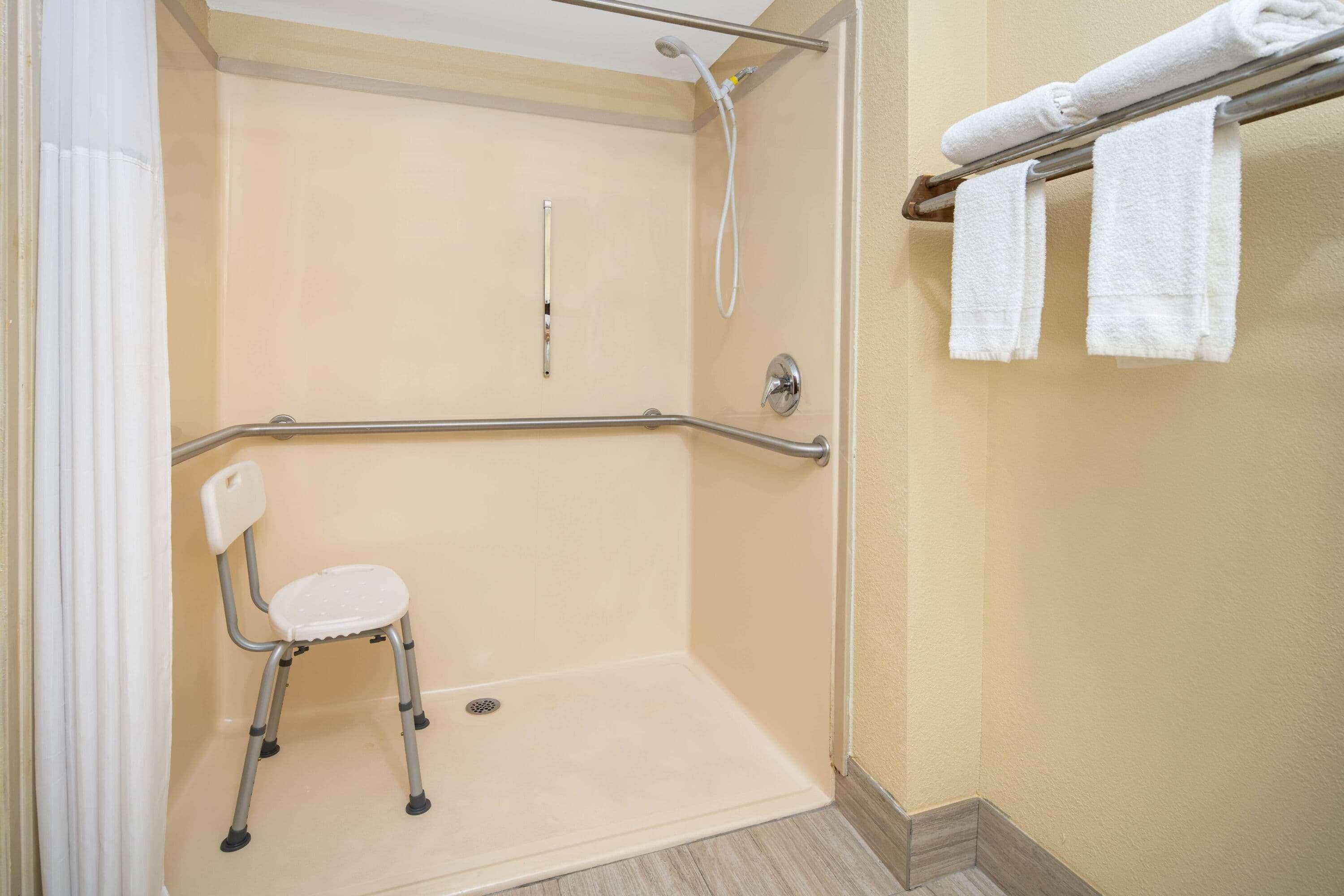 Una ducha accesible para sillas de ruedas con una barra de apoyo, una silla blanca y un perchero con toallas.