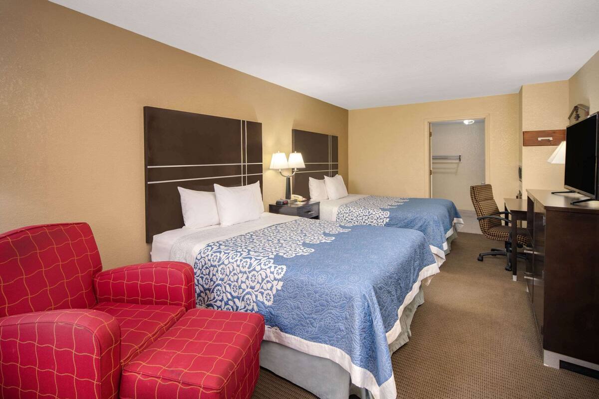 Ein Hotelzimmer mit zwei Betten, einem roten Sessel, einem Schreibtisch mit Fernseher und einem Stuhl.