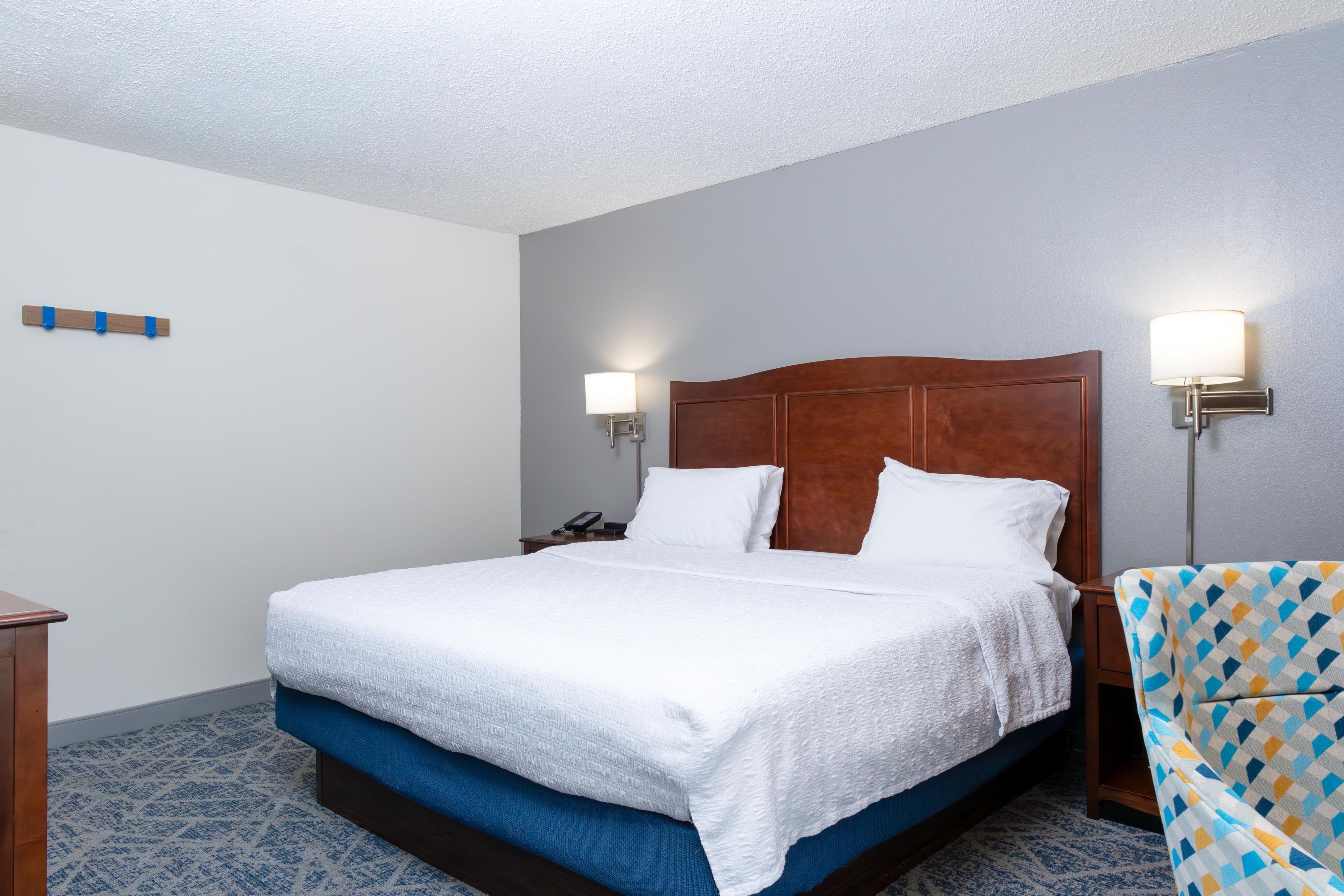A hotel room with a large bed, two bedside lamps, a telephone, and a patterned armchair.
