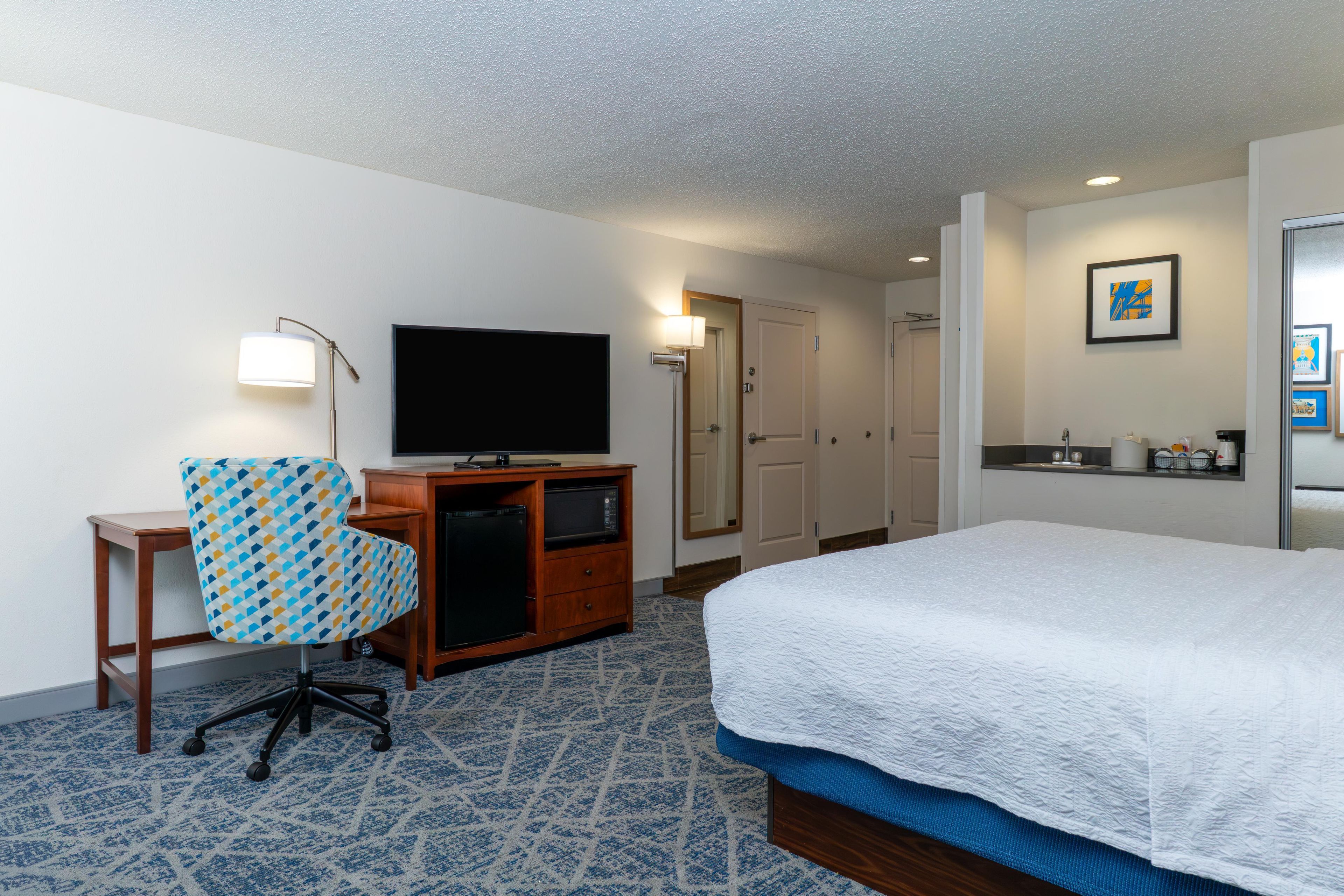 A hotel room with a bed, a desk with a television, a chair, a microwave, and a door.