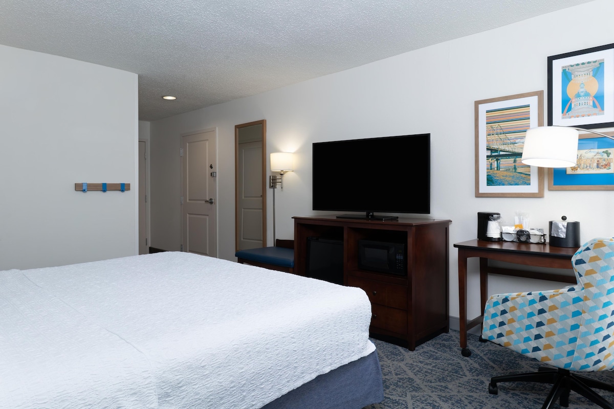 A hotel room with a bed, a television on a wooden stand, a desk with a lamp, and a chair.
