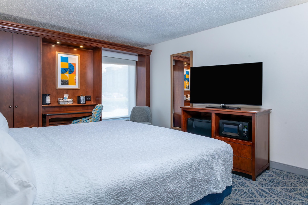 A hotel room with a large bed, a flat-screen TV, a desk with a chair, and a window.