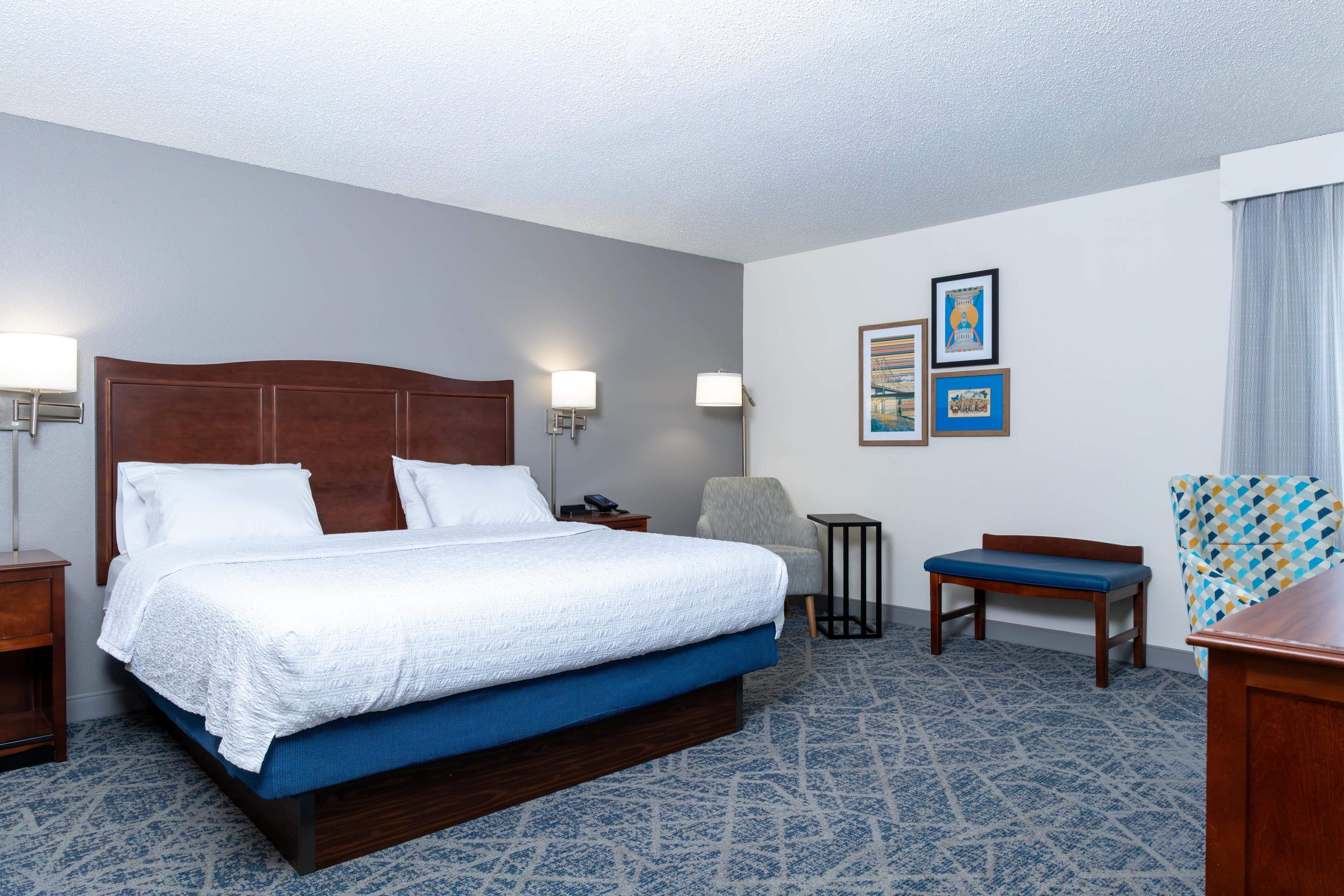 A hotel room with a large bed, two bedside lamps, a chair, a small table, and two framed pictures on the wall.