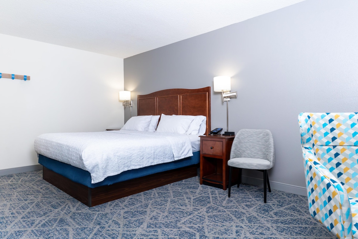 A hotel room with a bed, bedside table, chair, and a patterned carpet.