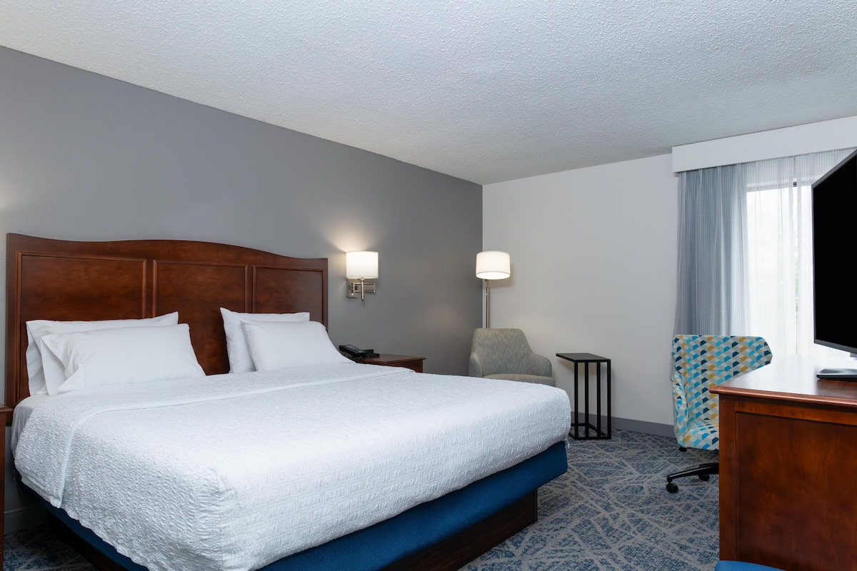 A hotel room with a large bed, a desk, a chair, a lamp, and a window with curtains.