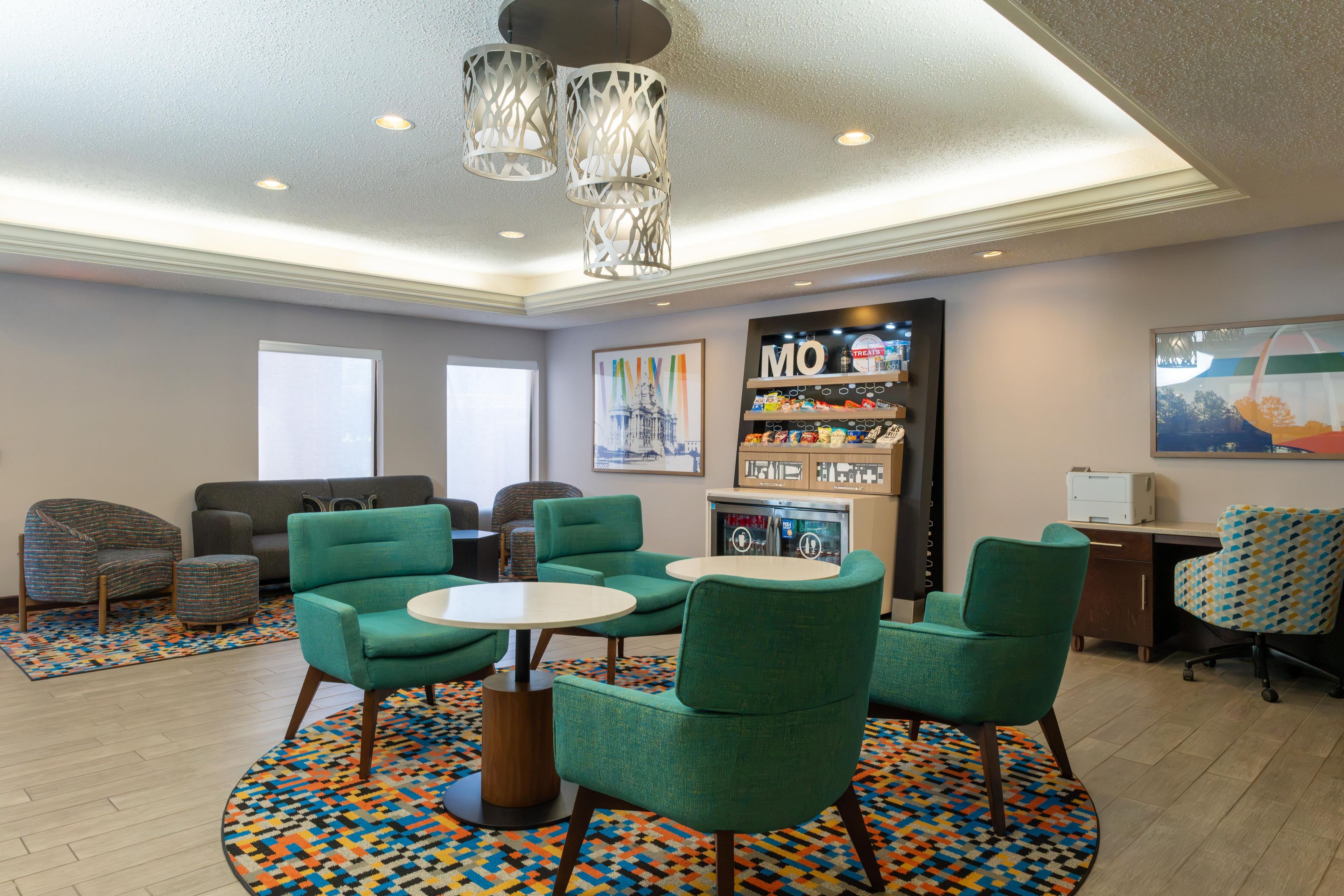 A modern lounge area with green upholstered chairs, a coffee table, a vending machine, and a wall-mounted refrigerator.