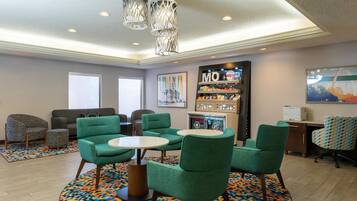 A modern lounge area with green upholstered chairs, a coffee table, a vending machine, and a wall-mounted refrigerator.