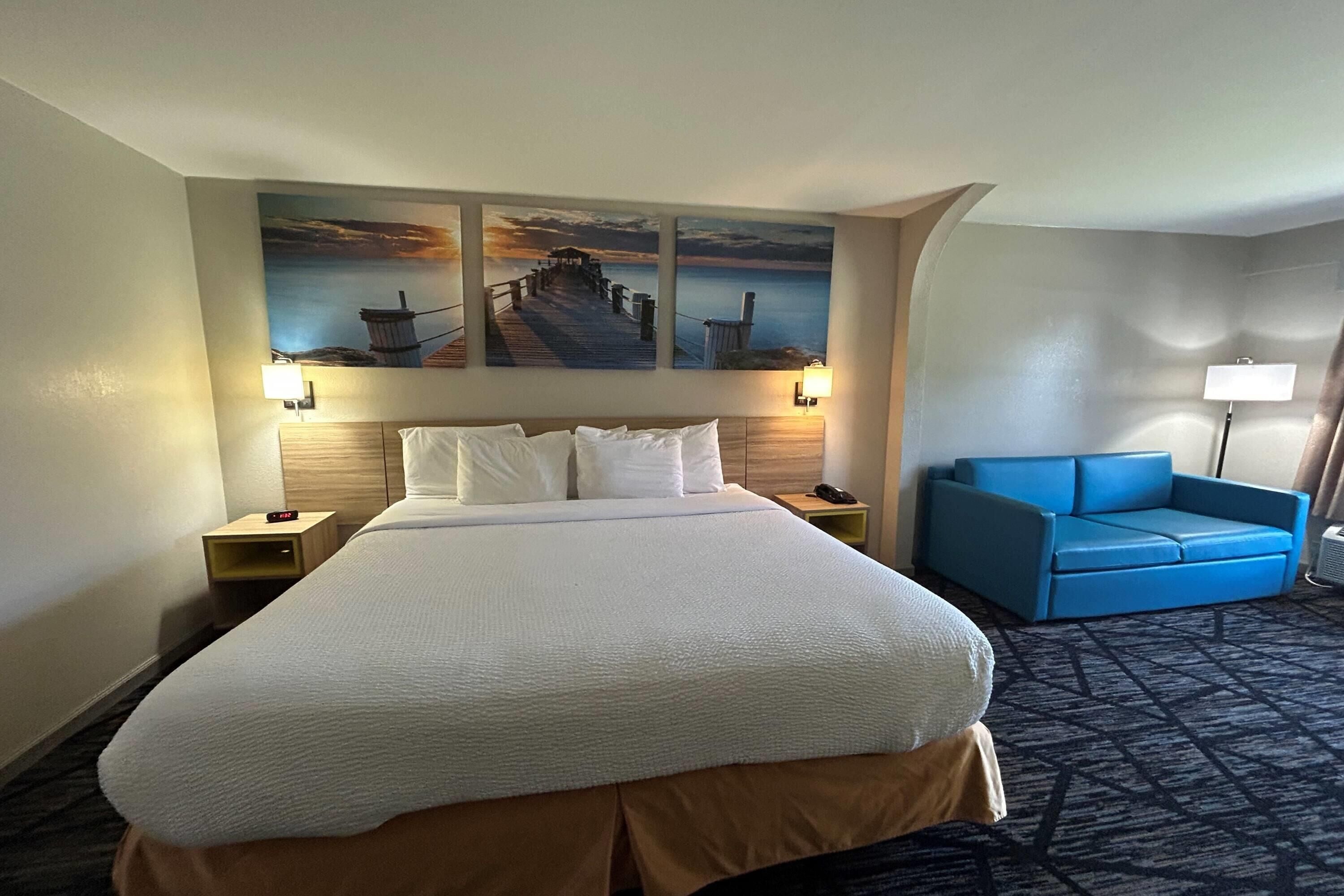 A hotel room with a large bed, a blue sofa, a painting of a pier, and a lamp.
