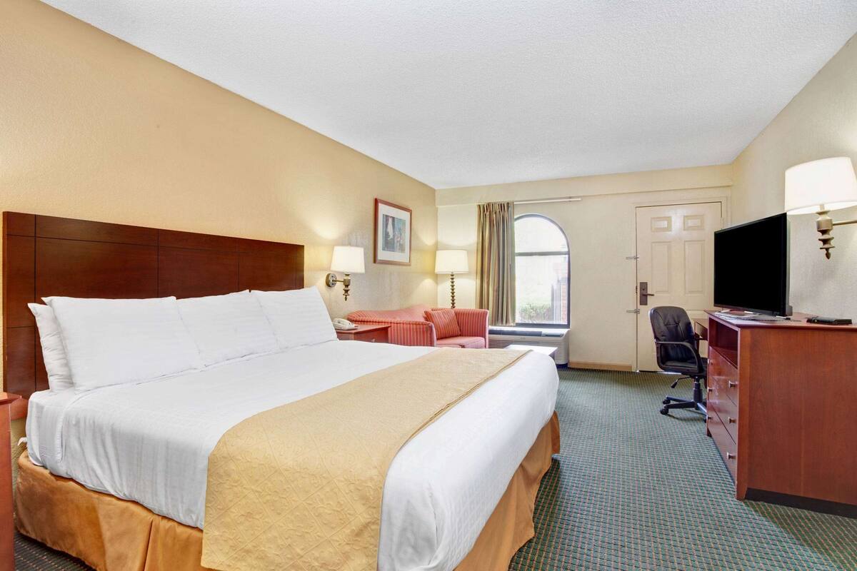 Ein Hotelzimmer mit einem großen Bett, einem Schreibtisch, einem Stuhl, einem Fernseher und einem Fenster mit Vorhängen.
