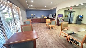 A hotel room with a wooden floor, a window with blinds, a kitchenette, a refrigerator, a vending machine, and a small table with chairs. - Days Inn & Suites by Wyndham Stockbridge South Atlanta (Stockbridge)