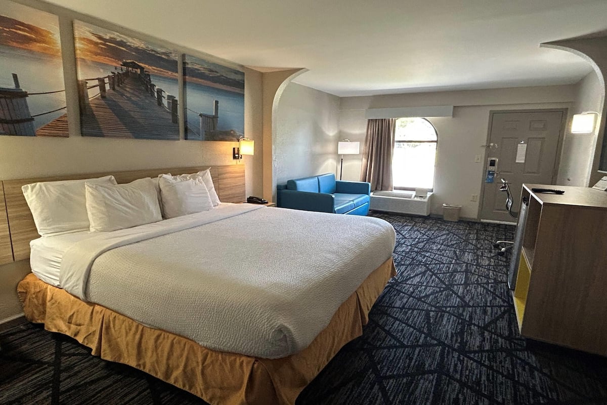 Ein Hotelzimmer mit einem großen Bett, einem blauen Sessel, einem Schreibtisch und einem Gemälde eines Piers.
