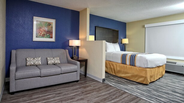 A hotel room with a gray sofa, a bed with a striped bedspread, a painting on the wall, and a projector screen. - Eagles Nest Inn (Statesboro)
