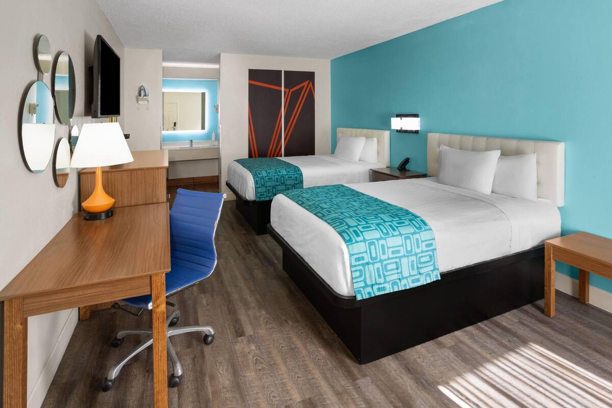 A hotel room with two beds, a desk, a chair, a TV, and a bathroom visible through a glass partition.