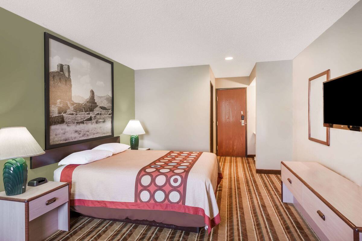 Une chambre d’hôtel avec un lit, une table de chevet avec une lampe, un téléviseur fixé au mur et un tableau encadré accroché au mur.