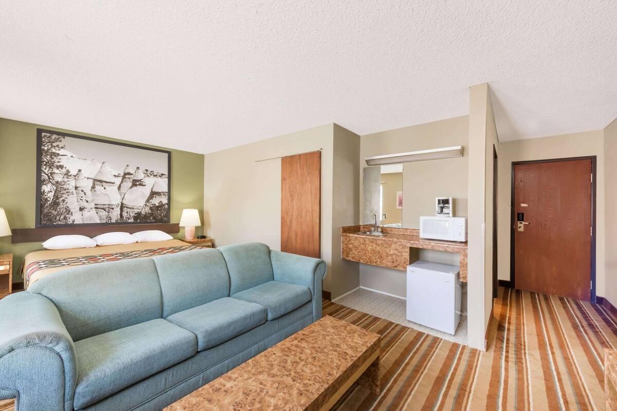 Une chambre d’hôtel avec un canapé bleu clair, un banc en bois, un lit orné d’un grand tableau encadré, une salle de bain avec un miroir, un lavabo et un petit réfrigérateur.