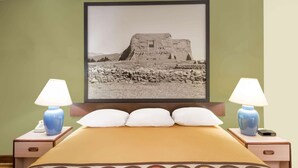 A hotel room with a bed, two bedside lamps, a framed picture of an ancient structure, and a nightstand with a phone.
