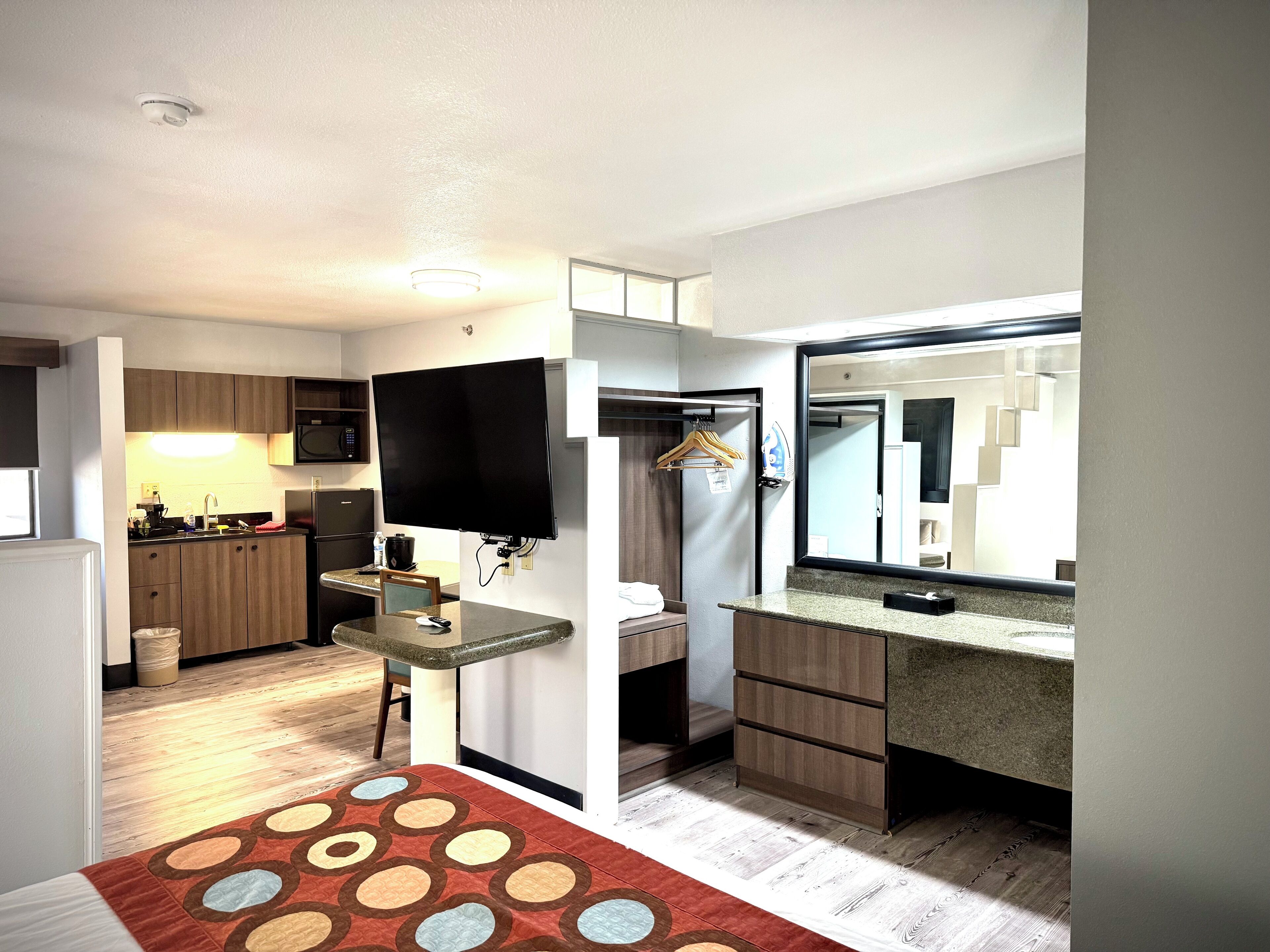 A compact hotel room with a kitchenette, a flat-screen TV, a mirror, and a small seating area.