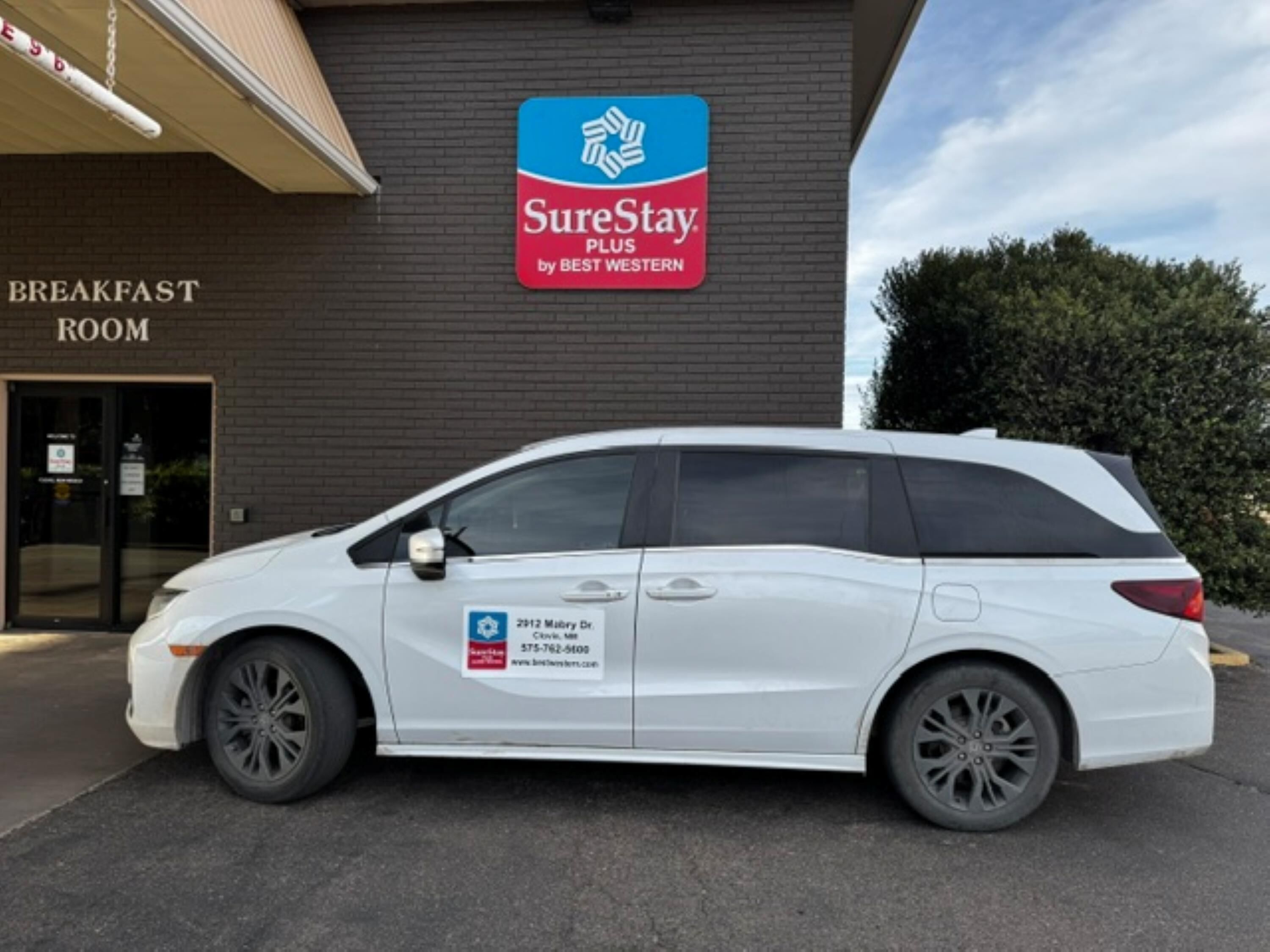 A white minivan parked in front of a building with a sign for SureStay Plus by Best Western.