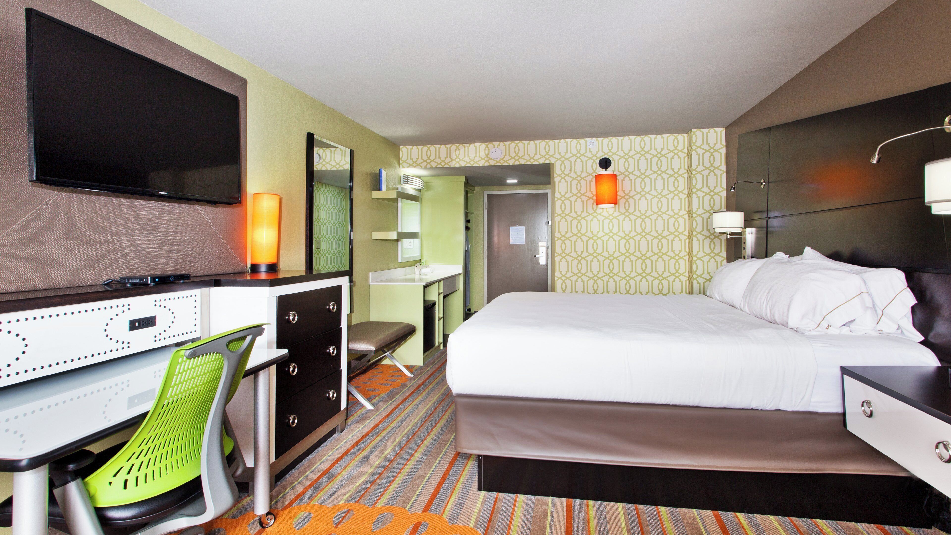 A hotel room with a bed, a desk with a chair, a TV, and a bathroom visible through an open door.