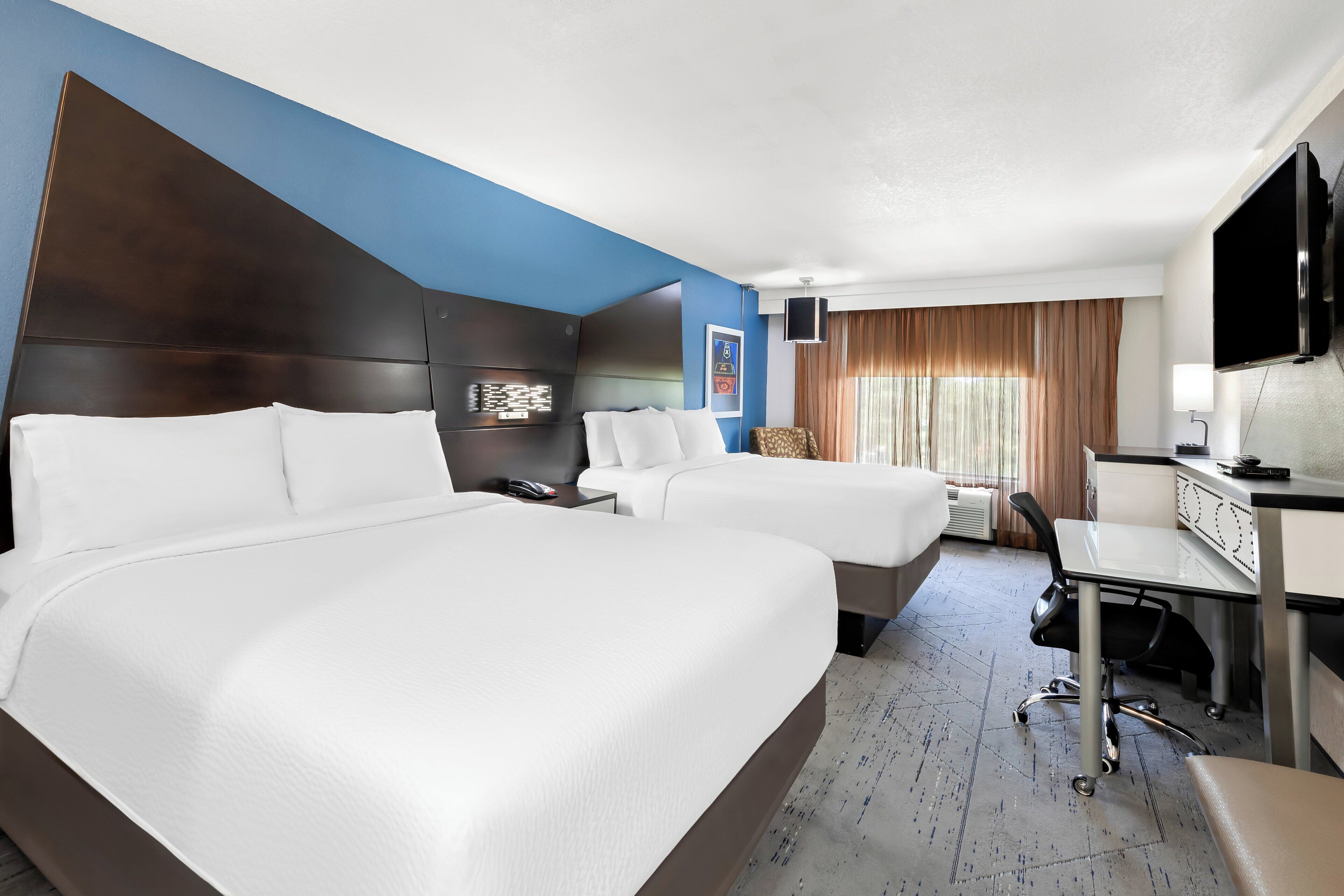 A hotel room with two beds, a desk, a chair, a television, and a window with curtains.