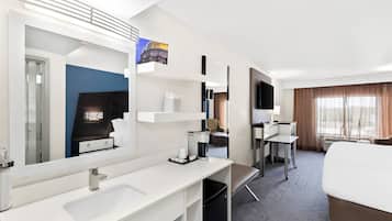 A hotel room with a large bed, a desk with a chair, a white vanity with a mirror, a small refrigerator, and a window with curtains.