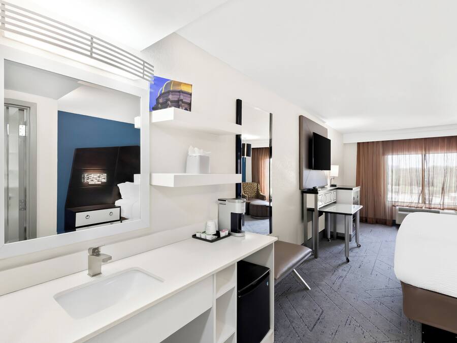 Habitación de hotel con una cama grande, un escritorio con silla, una tocador blanco con espejo, un refrigerador pequeño y una ventana con cortinas.