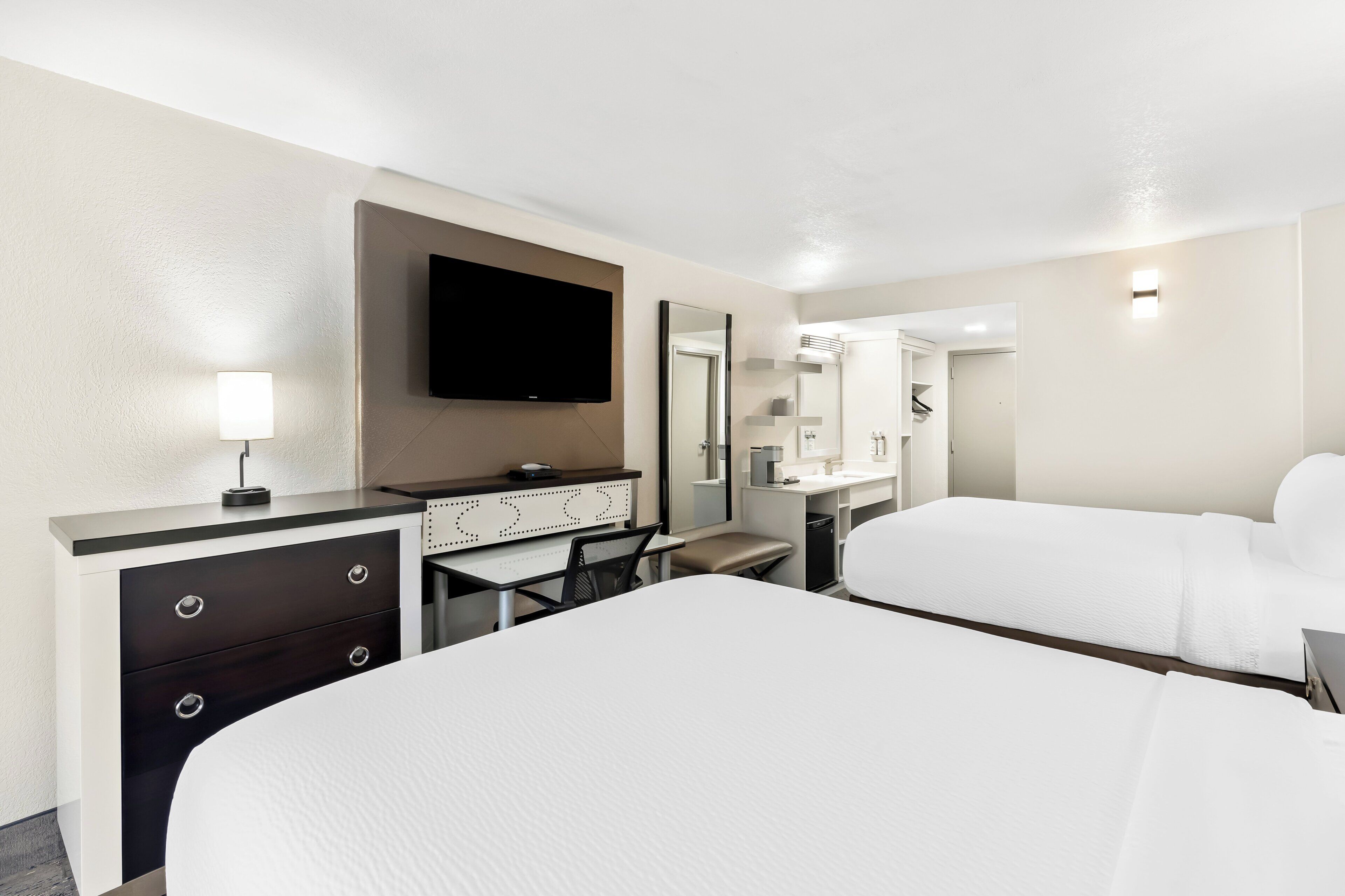 A hotel room with a large bed, a desk with drawers, a flat-screen TV, a mirror, and a bathroom visible through an open door.