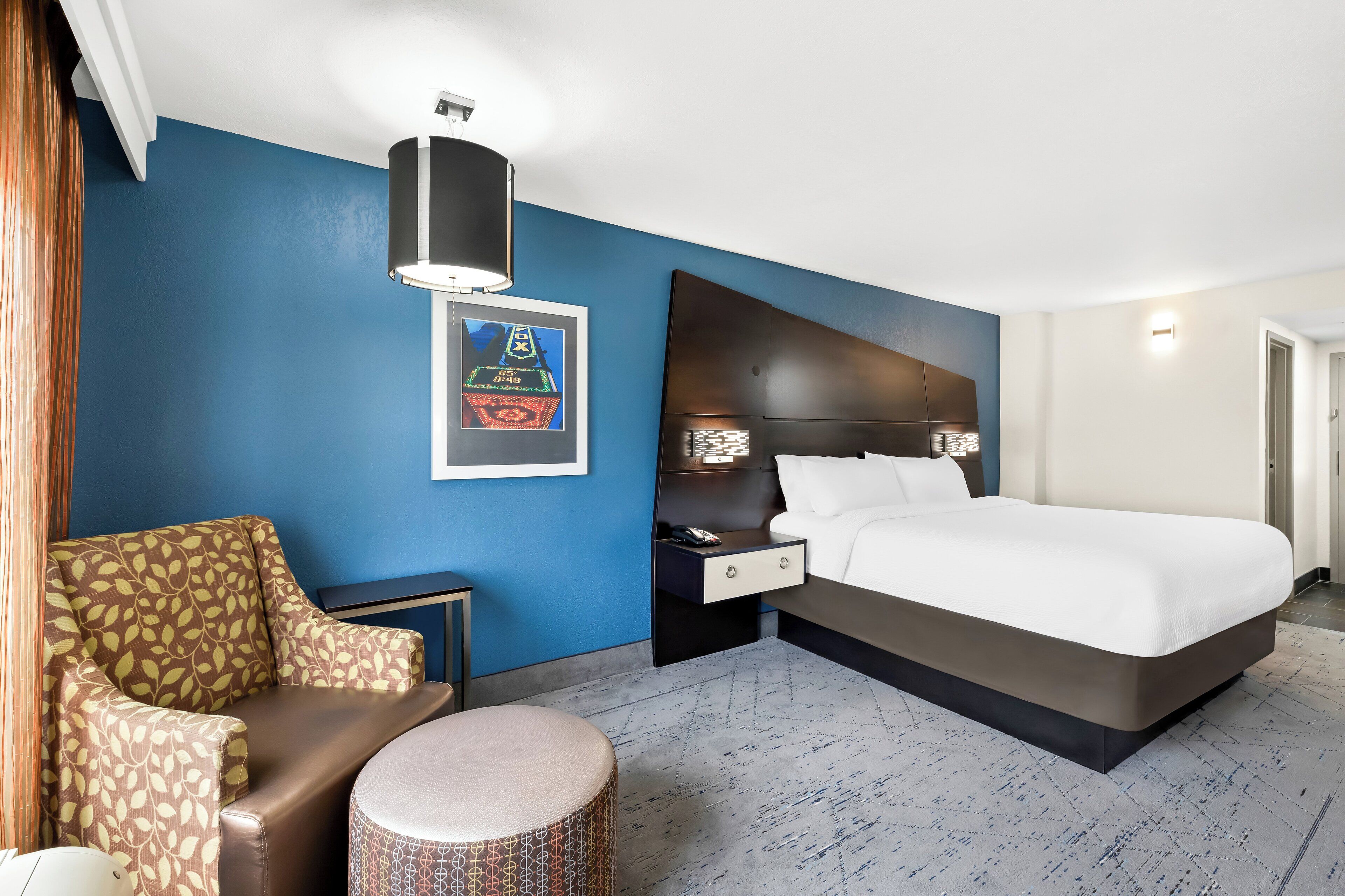 Ein Hotelzimmer mit einem großen Bett, einer Couch, einem kleinen runden Hocker, einem Nachttisch und einer Wandlampe.