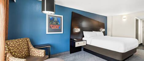 Una habitación de hotel con una cama grande, un sofá, un otomanillo redondo, una mesita de noche y una lámpara de pared.