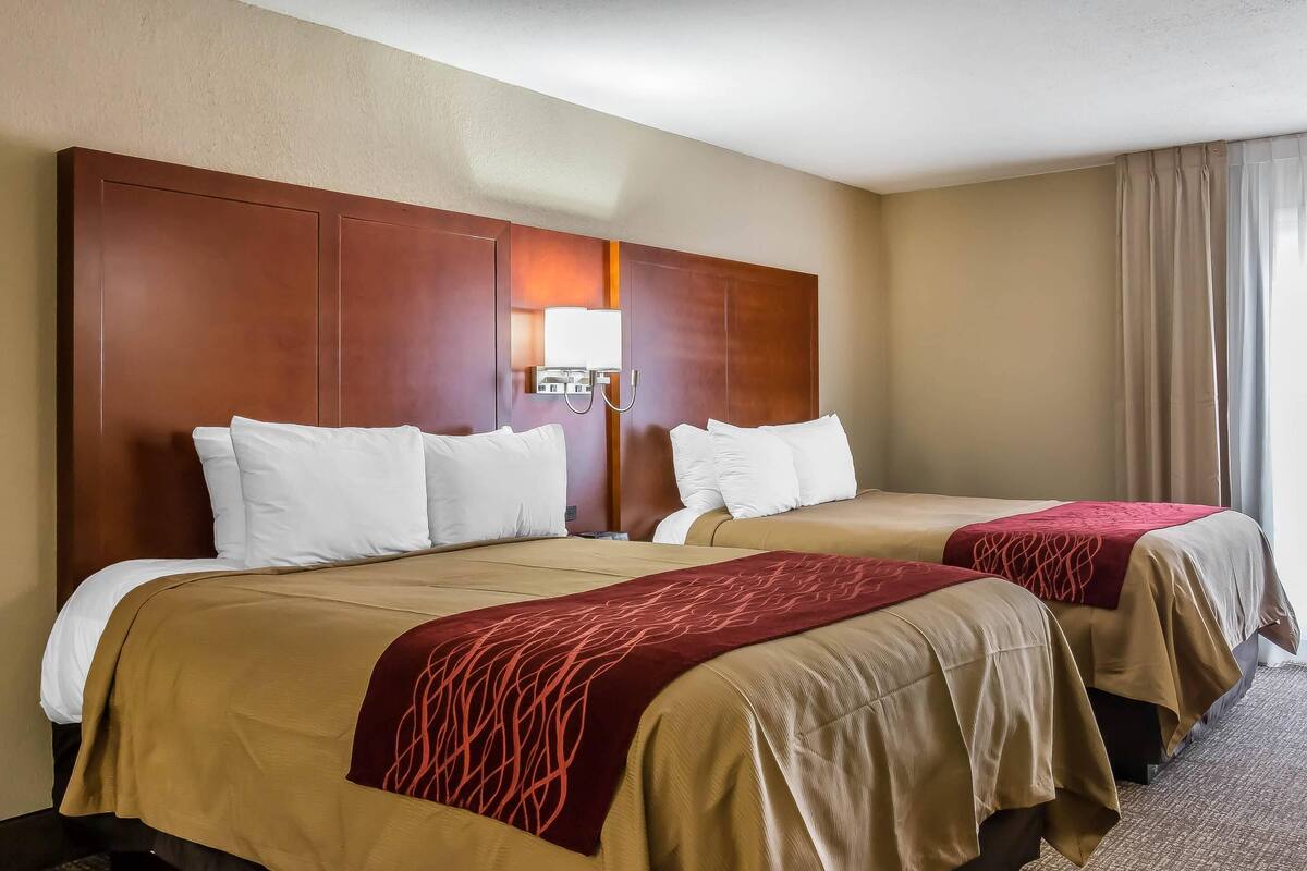 A hotel room with two beds, a wooden headboard, a nightstand, and a window with curtains.