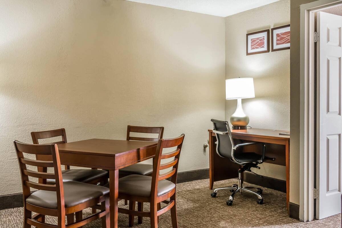A hotel room with a wooden dining table, four chairs, a desk with a chair, a lamp, and two framed pictures on the wall.