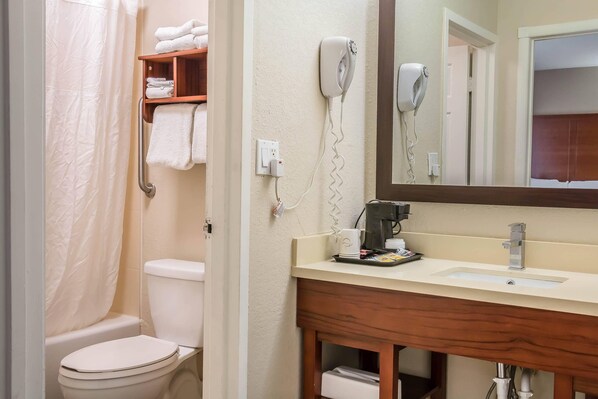 A bathroom with a toilet, a shower curtain, a towel rack with towels, a hairdryer, a coffee maker, and a sink with a mirror above it.