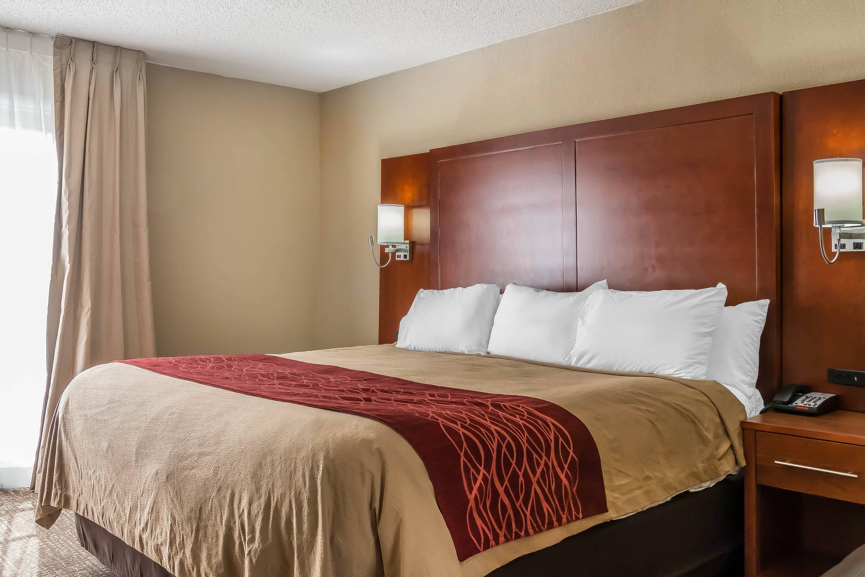 Quarto de hotel com uma cama grande, paredes bege, cabeceira de madeira, duas luminárias de cabeceira e um cobertor vermelho.