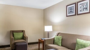 Une chambre d’hôtel avec un canapé beige, un fauteuil orné d’un coussin vert, une petite table avec une lampe et des tableaux encadrés accrochés au mur.