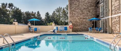 En swimmingpool med blå kakelplattor, omgiven av solstolar och parasoller, med en byggnad i tegel som syns i bakgrunden.