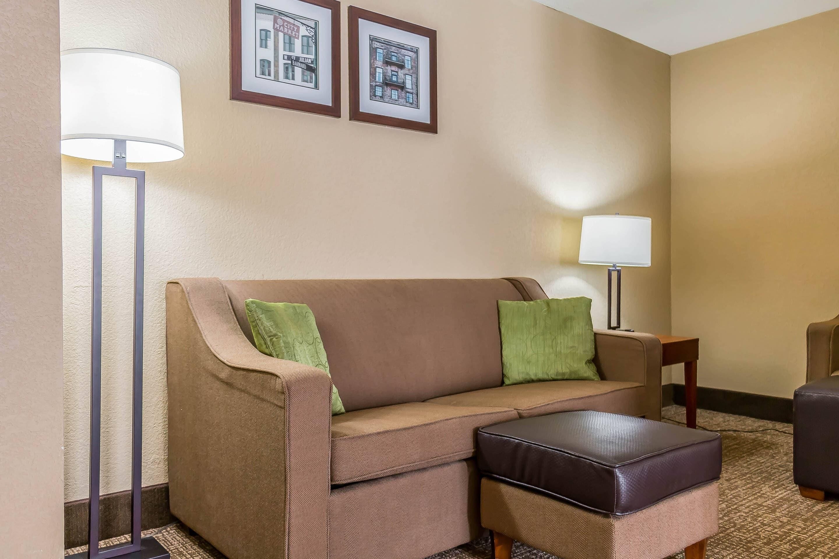 A hotel room with a brown sofa, a matching ottoman, a floor lamp, a side table, and two framed pictures on the wall.