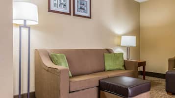 A hotel room with a brown sofa, a matching ottoman, a floor lamp, a side table, and two framed pictures on the wall.