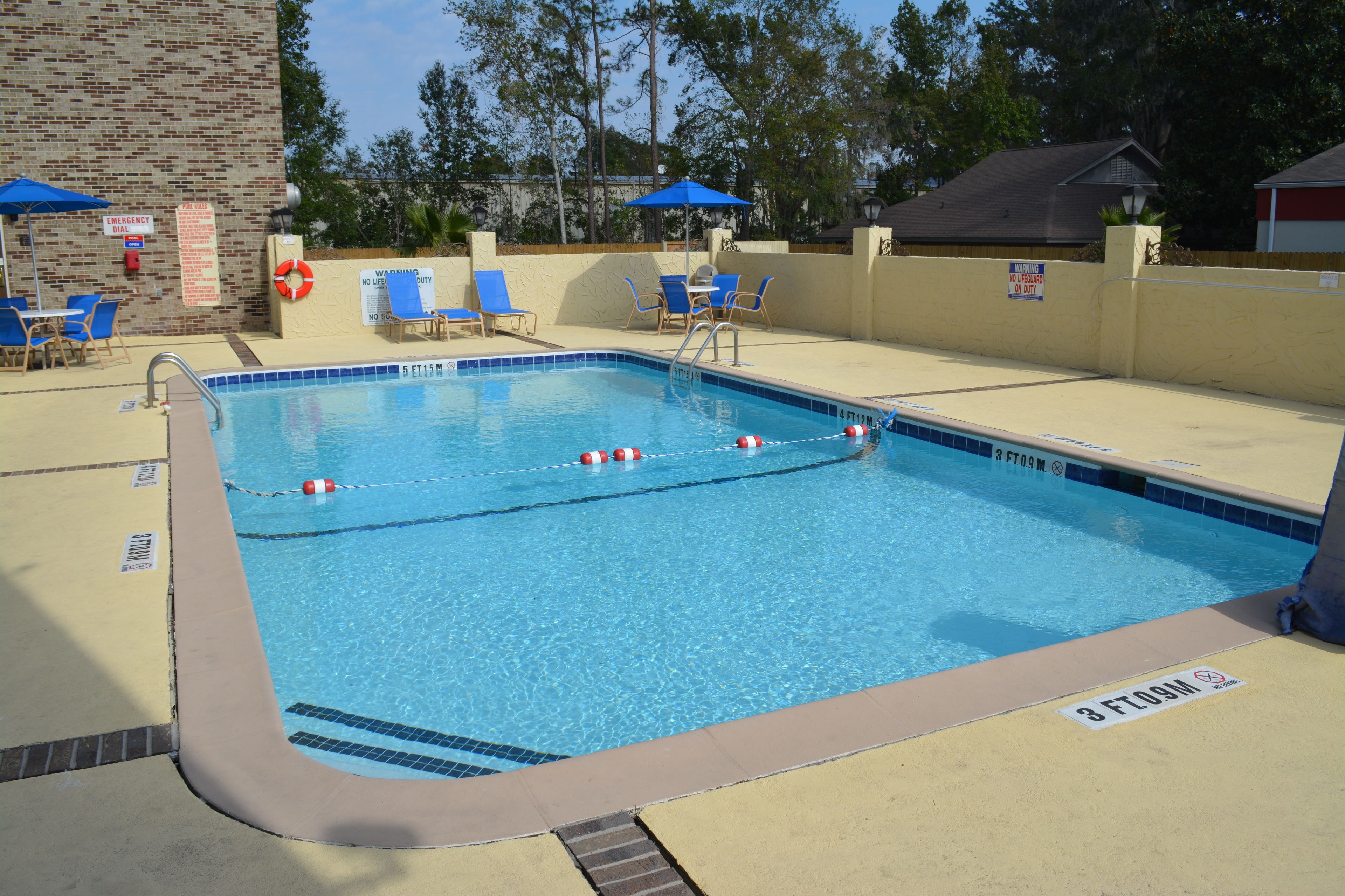 Una piscina rectangular con borde azul, rodeada por un deck color beige. Se observan sillas de descanso y sombrillas, además de un muro de ladrillo con un flotador salvavidas y un cartel de llamada de emergencia.