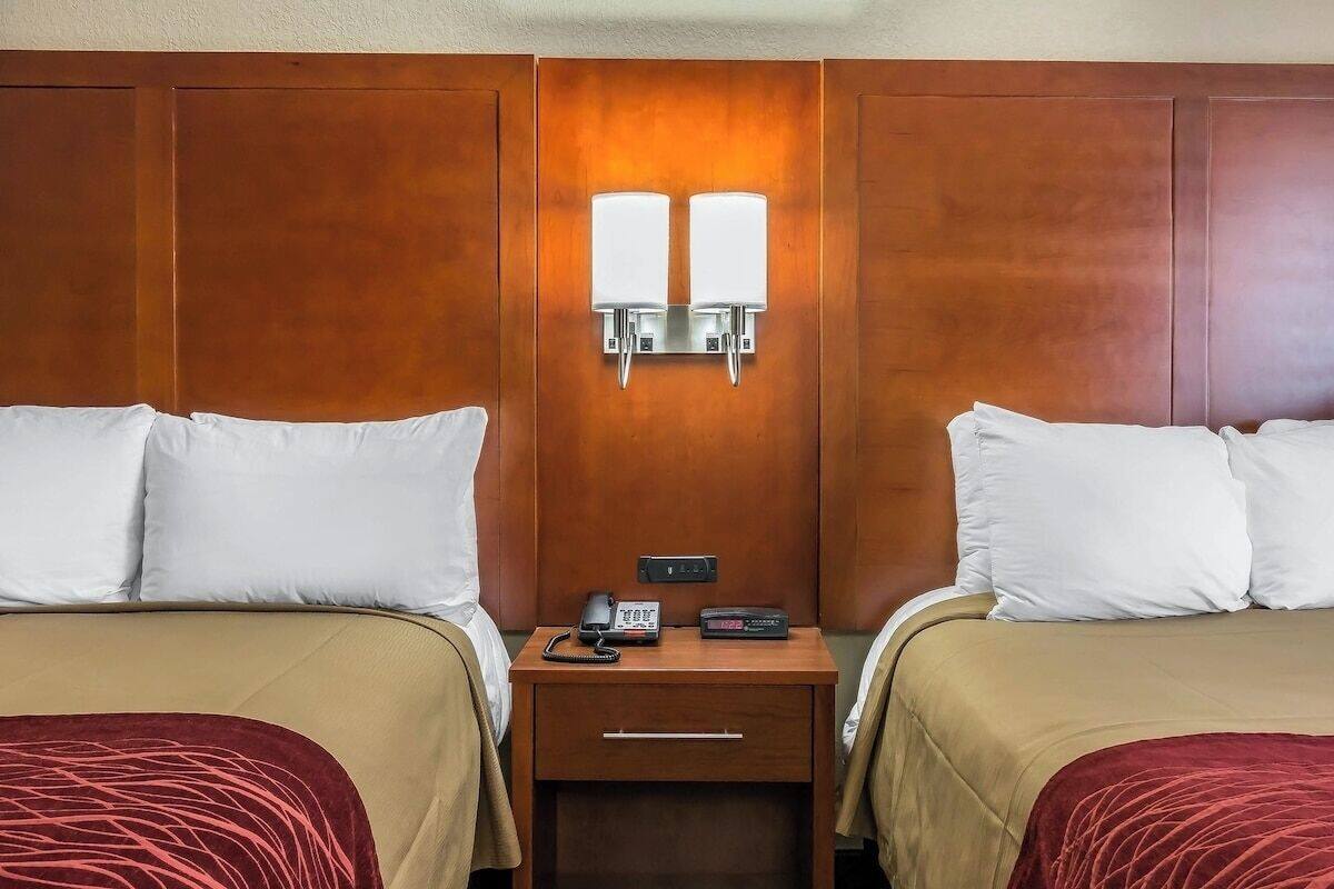 A hotel room with two beds, a nightstand with a phone and a clock, and a wall-mounted light fixture.