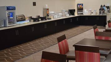 A hotel dining area with a buffet setup, including a refrigerator, coffee machines, and a variety of food containers.