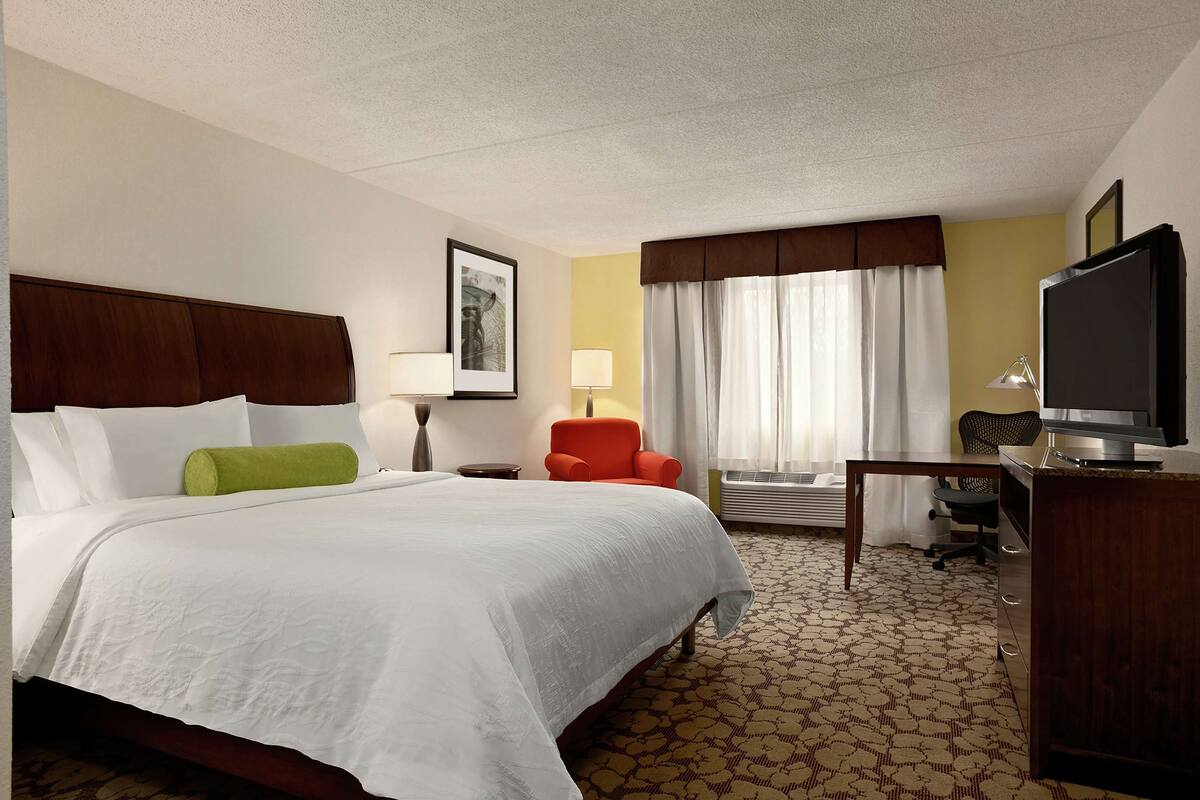 A hotel room with a large bed, a desk with a television, a chair, and a window with curtains.