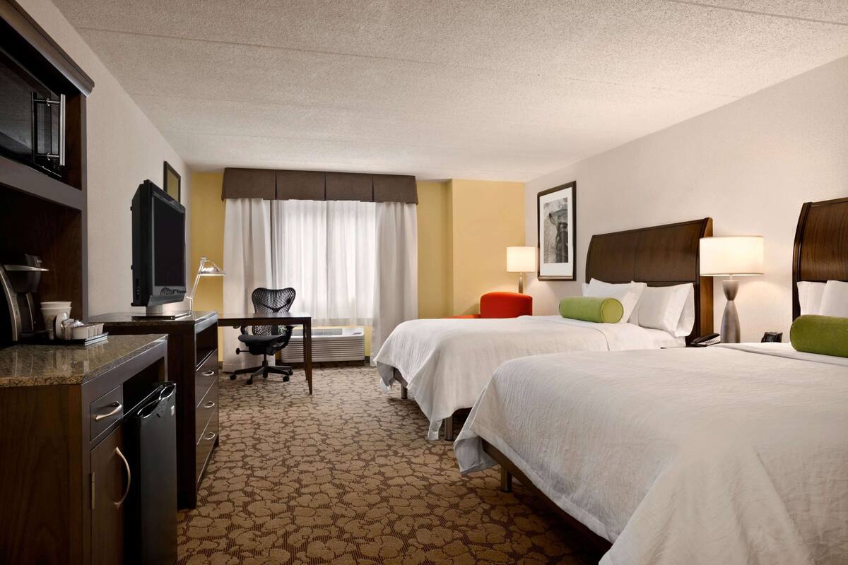 A hotel room with two beds, a desk with a chair, a television, and a window with curtains.