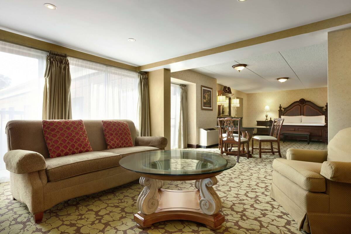 A hotel room with a sofa, armchair, and a glass-top coffee table. There is a dining area with a table and chairs.