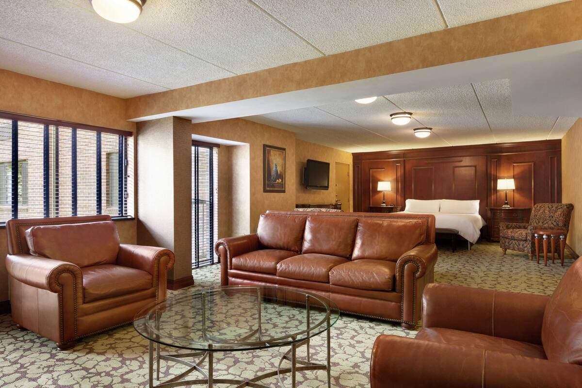 A hotel room with a brown leather sofa, a glass coffee table, a bed, and a TV mounted on the wall.
