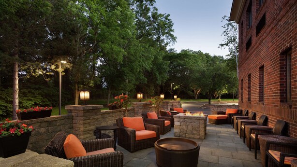An outdoor patio area with wicker furniture, stone planters with flowers, and a brick building in the background. - Hilton Garden Inn Rochester/Pittsford (Pittsford)